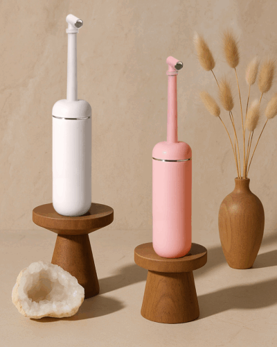 Roze en witte draagbare bidetflessen op houten standaarden in een serene Japandi setting, perfect voor persoonlijke hygiëne onderweg.